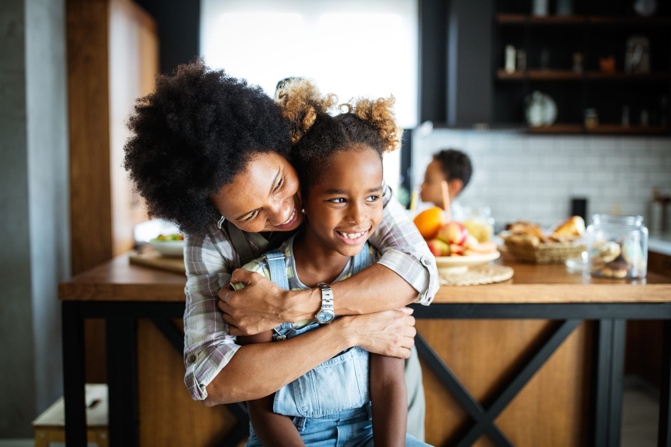 A mother laughs while embracing her daughter, who smiles warmly, in a bright kitchen